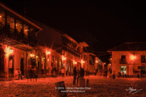 Colombia, South America - Tourists And Some Local Residents On The Eastern Corner Of The Plaza Mayor In The Historic 16th Century Town of Villa de Leyva In The Boyacá Department, Just After Sunset; Night Shot Lit By Street Lights